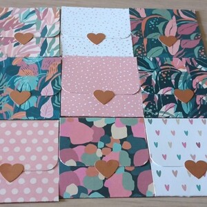 Peut inclure: Un ensemble de neuf enveloppes carrées en papier disposées en grille, chacune présentant un motif unique incluant des fleurs, des pois et des cœurs. Chaque enveloppe est fermée par une petite découpe en papier marron en forme de cœur.