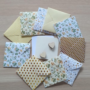 Peut inclure: Un ensemble d'enveloppes carrées en papier présentant divers motifs, notamment des alvéoles jaunes, des imprimés floraux et des abeilles, disposées en cercle autour d'une carte blanche centrale ornée de deux petits cœurs en bois.