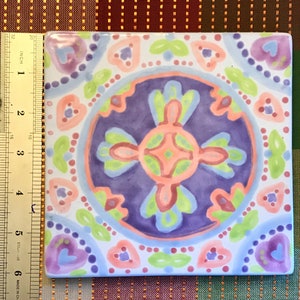 May include: A square ceramic tile with a colorful, intricate design. The tile features a central circle with a cross-shaped pattern in shades of pink, green, and blue. The circle is surrounded by a border of smaller circles and hearts in various colors.