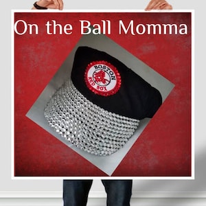 May include: A black baseball cap with a red and white Boston Red Sox logo and a silver rhinestone trim. The text "On the Ball Momma" is written above the cap.