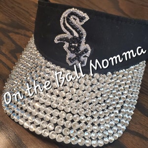 May include: Black visor with a white and black embroidered baseball logo and the text "On the Ball Momma" on the brim. The visor is covered in clear rhinestones.