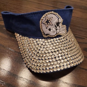 May include: A navy blue visor with a silver star and helmet design. The visor is covered in clear rhinestones.