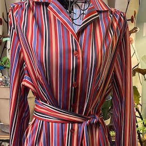May include: A long-sleeved button-down shirt with a striped pattern in red, blue, white, and black. The shirt has a tie at the waist.