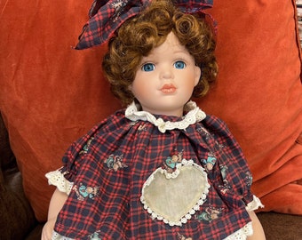 May include: A porcelain doll with brown curly hair wearing a red and black plaid dress with a white heart applique and reindeer print. The doll is wearing brown reindeer-shaped shoes.