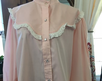 May include: A light pink blouse with a high collar and long sleeves. The blouse features a decorative yoke with white lace trim and silver snap closures down the front. The cuffs also have lace trim.