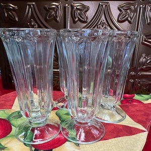 May include: Set of four clear glass sundae or dessert glasses. Each glass has a flared, ribbed design and sits on a round, clear base. The glasses are arranged on a red and yellow patterned tablecloth with apple designs.