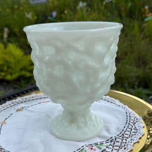 Könnte beinhalten: Ein weißes, strukturiertes Glasbecher mit einem Sockel. Der Becher steht auf einer weißen Tischdecke mit Blumenstickerei, die auf einem goldfarbenen Tablett platziert ist. Der Hintergrund ist mit Grün verschwommen.