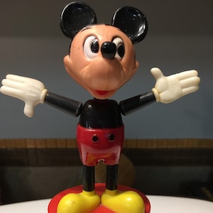 May include: A red, black, and yellow Mickey Mouse puppet with a push button base. The puppet is made by Kohner and is labeled "Mickey Mouse Maxi-Puppet".