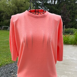 May include: A coral pink short-sleeved sweater with a round neckline. The sweater is made of a soft, lightweight material and has a slightly cropped fit.