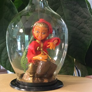 May include: A clear glass dome with a black base encloses a small plastic figurine of a person wearing a red hooded cloak and holding a brown drum. The figurine is sitting on a small rock with green foliage.