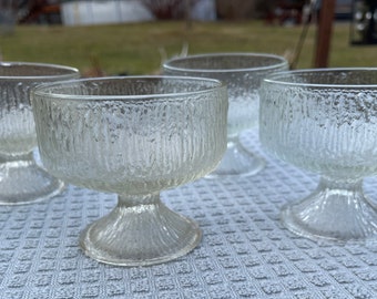 May include: Four clear glass dessert bowls with a textured, ribbed design. The bowls have a pedestal base and are arranged on a gray and white patterned surface.