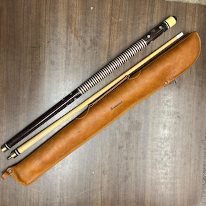 May include: A two-piece pool cue and a tan leather carrying case. The cue features a dark brown butt with a white and brown striped wrap section, white inlays, and a light-colored wooden shaft. The case is embossed with the word Brunswick.