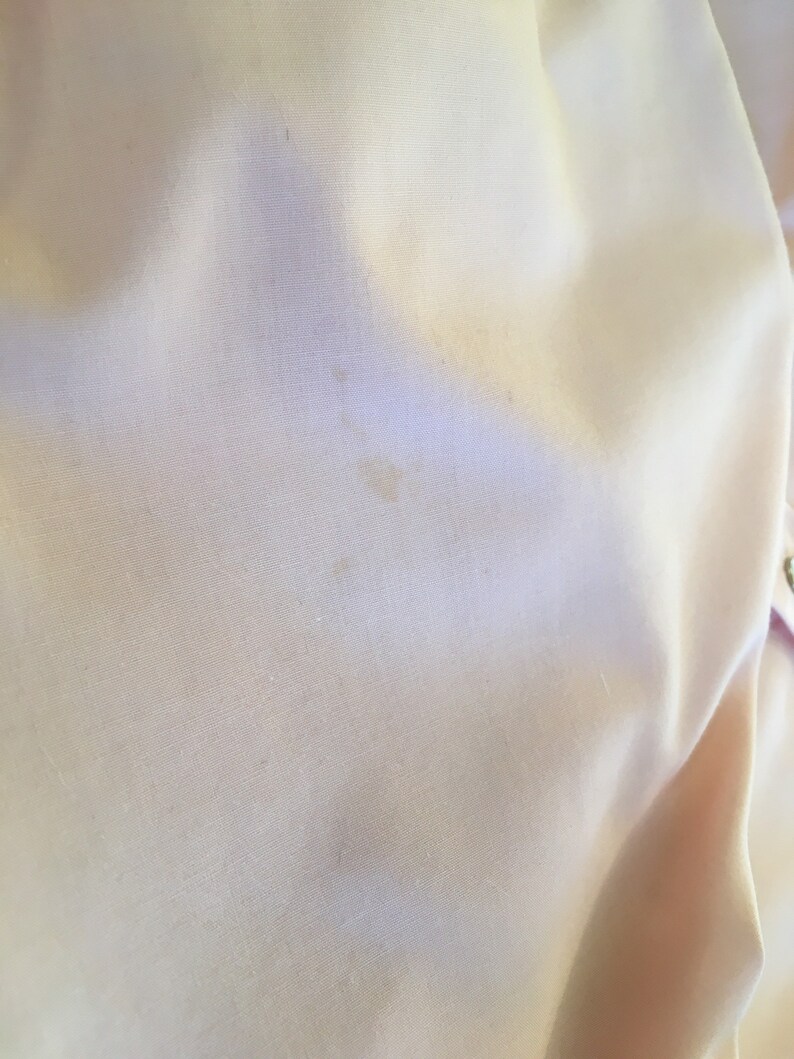May include: Close-up of a pale yellow fabric with subtle texture and slight discoloration. The fabric appears to be smooth and possibly part of a garment or textile item. The image highlights the material's color and surface details.