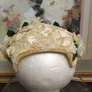 May include: A white vintage hat with a floral design. The hat is made of fabric and has a velvet trim. The hat is decorated with white flowers and green leaves.