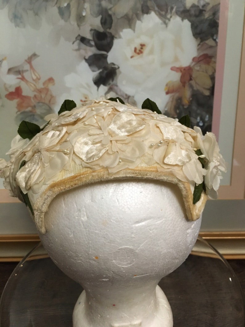 May include: A white vintage hat with a floral design. The hat is made of fabric and has a velvet trim. The hat is decorated with white flowers and green leaves.