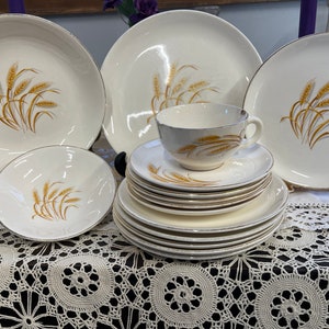 May include: A vintage cream-colored dinnerware set featuring a wheat design. The set includes plates, bowls, saucers, and a teacup. Each piece has a gold trim. The wheat design is in shades of gold and brown.