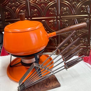 May include: A bright orange fondue pot with a wooden handle and lid, resting on a black metal stand with a burner. Beside it is a set of six metal fondue forks with wooden handles, organized in a wooden block stand with a metal support frame.