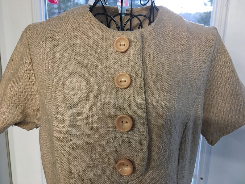 Puede incluir: Un vestido de lino beige con mangas cortas y una fila de cuatro botones marrones claros en la parte delantera.