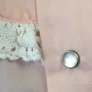 May include: Close-up of a light pink garment with a white crocheted lace collar. A silver button with a pearlescent center is visible. The fabric has vertical stitching details.