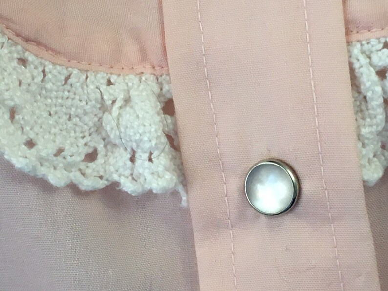 May include: Close-up of a light pink garment with a white crocheted lace collar. A silver button with a pearlescent center is visible. The fabric has vertical stitching details.