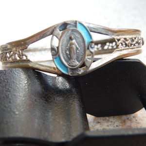 May include: Silver bracelet with a blue and white enamel oval charm featuring a depiction of the Virgin Mary. The bracelet has a delicate filigree design.