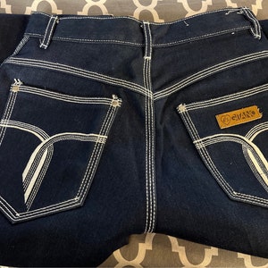 May include: A pair of dark blue denim jeans with white stitching. The back pockets have a unique design with white stitching that resembles a stylized letter 'Y'. A brown leather patch with the word "GIVANO" is sewn onto the right back pocket.