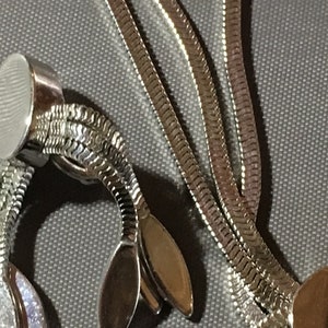 May include: A silver chain necklace with a round clasp and a decorative end piece with three oval shapes. The chain is made of a snake-like pattern.