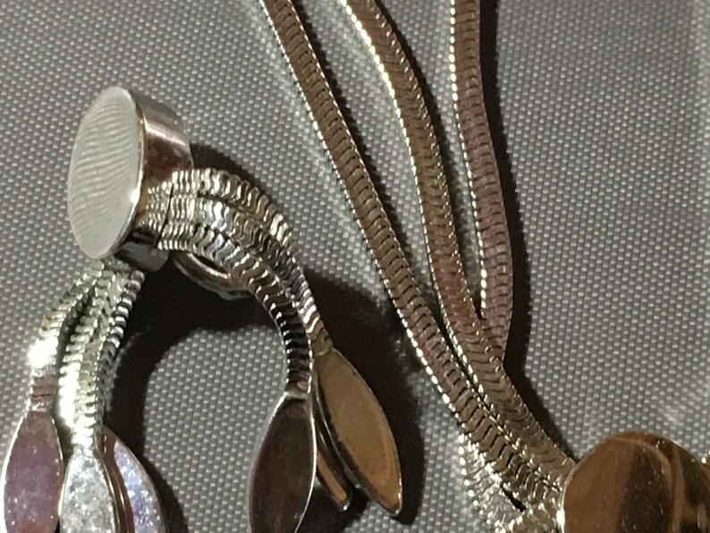 May include: A silver chain necklace with a round clasp and a decorative end piece with three oval shapes. The chain is made of a snake-like pattern.