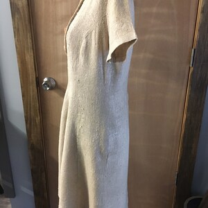 Puede incluir: Un vestido de lino beige con mangas cortas y cierre de botones en la parte delantera. El vestido tiene un corpi&ntilde;o ajustado y una falda ligeramente acampanada.