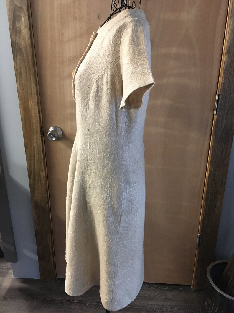 Puede incluir: Un vestido de lino beige con mangas cortas y cierre de botones en la parte delantera. El vestido tiene un corpi&ntilde;o ajustado y una falda ligeramente acampanada.