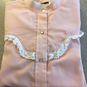 May include: A light pink button-down shirt with a stand-up collar and white lace trim. The shirt has a series of buttons down the front and on the cuffs. The shirt is folded neatly on a light gray surface.