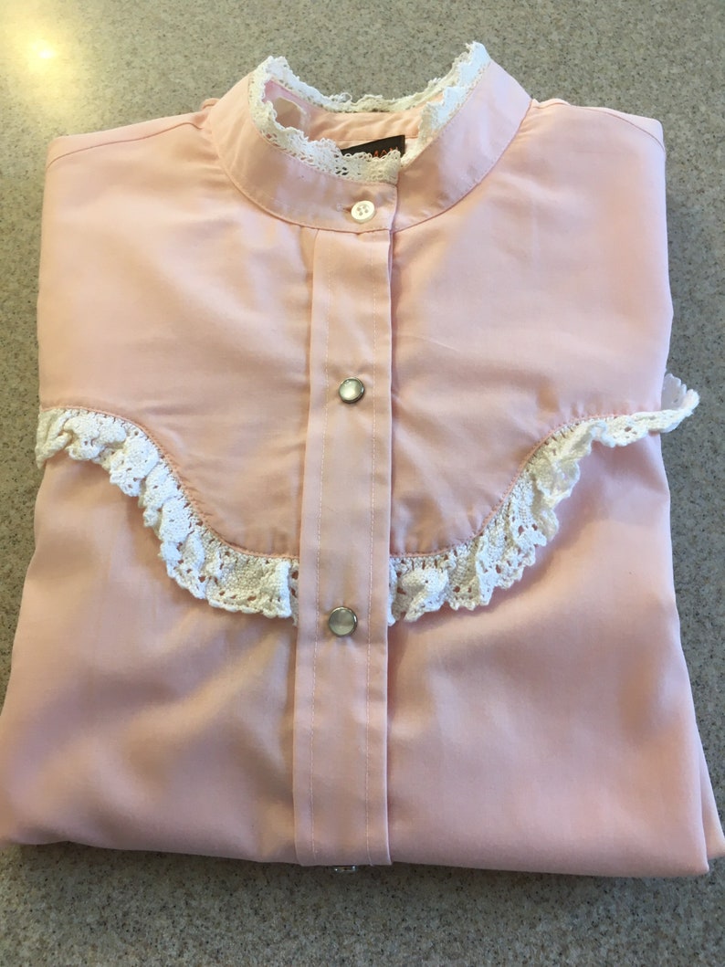 May include: A light pink button-down shirt with a stand-up collar and white lace trim. The shirt has a series of buttons down the front and on the cuffs. The shirt is folded neatly on a light gray surface.