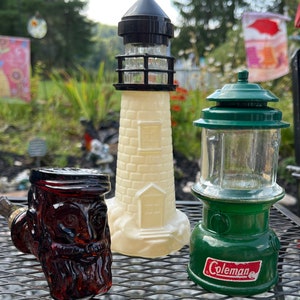 May include: Three vintage glass bottles shaped like a lighthouse, a lantern, and a pipe. The lighthouse bottle is white with a black top and a clear glass window. The lantern bottle is green with a clear glass window and a red label that says "Coleman". The pipe bottle is brown with a red glass face and beard.