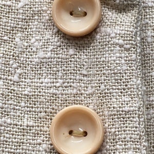 Puede incluir: Dos botones beige con un solo agujero en el centro, cosidos sobre una tela beige con un patr&oacute;n texturizado.