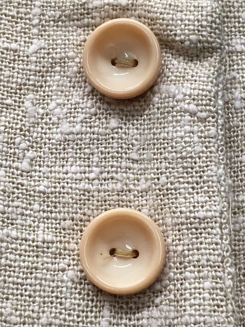 Puede incluir: Dos botones beige con un solo agujero en el centro, cosidos sobre una tela beige con un patr&oacute;n texturizado.