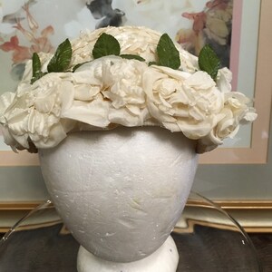 May include: A white hat made of fabric roses with green leaves around the brim.