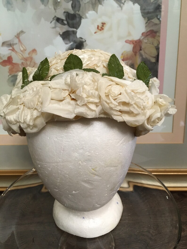 May include: A white hat made of fabric roses with green leaves around the brim.