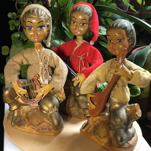 May include: Three vintage elf figurines playing musical instruments. The elf on the left is playing a lyre, the elf in the middle is playing a flute, and the elf on the right is playing a lute. The elves are wearing brown and gold clothing and have painted faces.