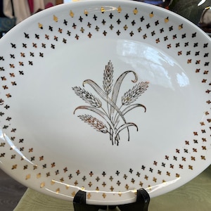 May include: Oval white serving platter with a gold wheat stalk design in the center. The rim is decorated with gold fleur-de-lis shapes. The platter is on a black stand, suitable for serving food.