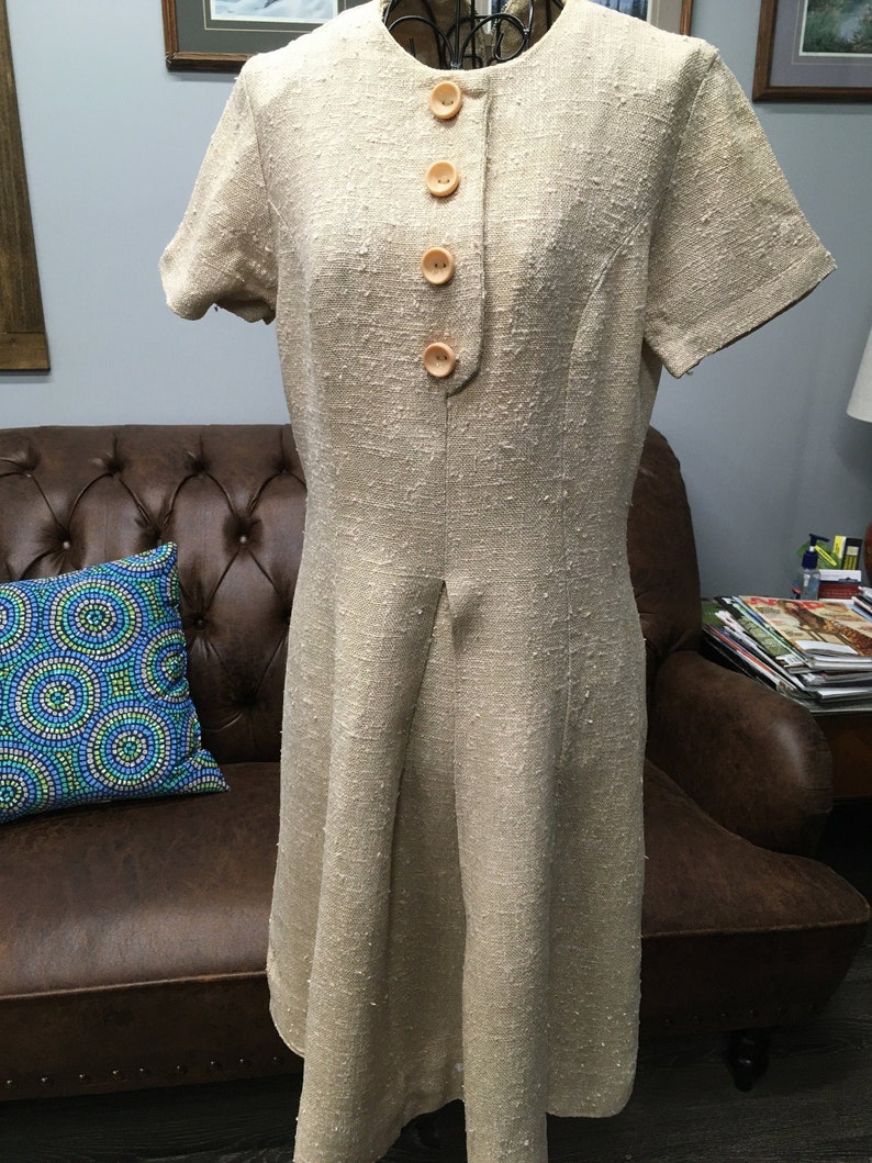 Puede incluir: Un vestido de lino beige con mangas cortas y cierre de botones en la parte delantera. El vestido tiene una silueta ligeramente acampanada y una tela texturizada.