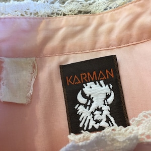 May include: Close-up of a pink garment with a white lace trim. The label reads "KARMAN" in orange lettering on a brown background, with a white embroidered design. A white fabric tag is also visible.