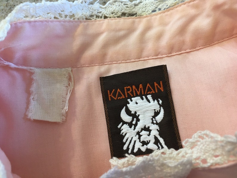 May include: Close-up of a pink garment with a white lace trim. The label reads "KARMAN" in orange lettering on a brown background, with a white embroidered design. A white fabric tag is also visible.