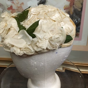 May include: A white fabric hat with a floral design. The hat is made of white fabric and is decorated with white fabric flowers. The hat is sitting on a white styrofoam head.