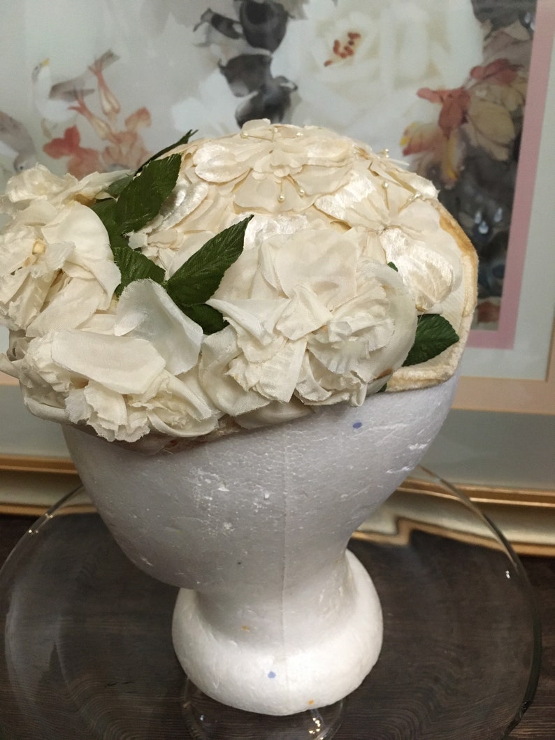 May include: A white fabric hat with a floral design. The hat is made of white fabric and is decorated with white fabric flowers. The hat is sitting on a white styrofoam head.