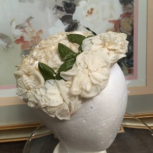 May include: A white hat made of fabric roses with green leaves. The hat is on a white mannequin head.