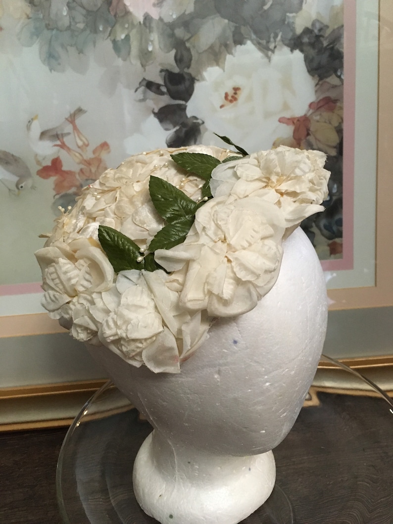 May include: A white hat made of fabric roses with green leaves. The hat is on a white mannequin head.