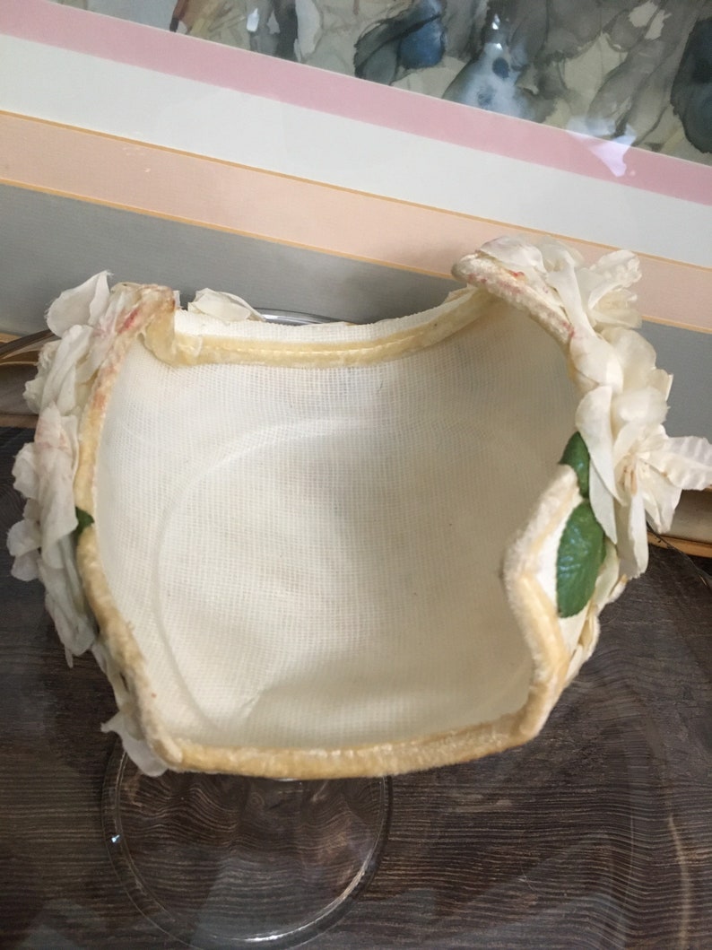 May include: A white fabric hat with a cream colored velvet trim and white fabric flowers with green leaves.