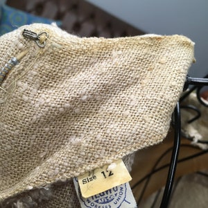 Puede incluir: Primer plano de una tela de lino beige con un patr&oacute;n de lunares blancos. Una etiqueta amarilla con el texto "Talla 12" est&aacute; unida a la tela. Una etiqueta azul y blanca con el texto "ILGWU AFL-CIO WORKERS UNION" tambi&eacute;n est&aacute; unida a la tela.