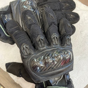 以下が含まれることがあります： カーボンファイバー製のナックルプロテクターとメッシュデザインを備えた、黒い革のオートバイ用グローブ。グローブは手首にベルクロ留めと星形のロゴが付いています。