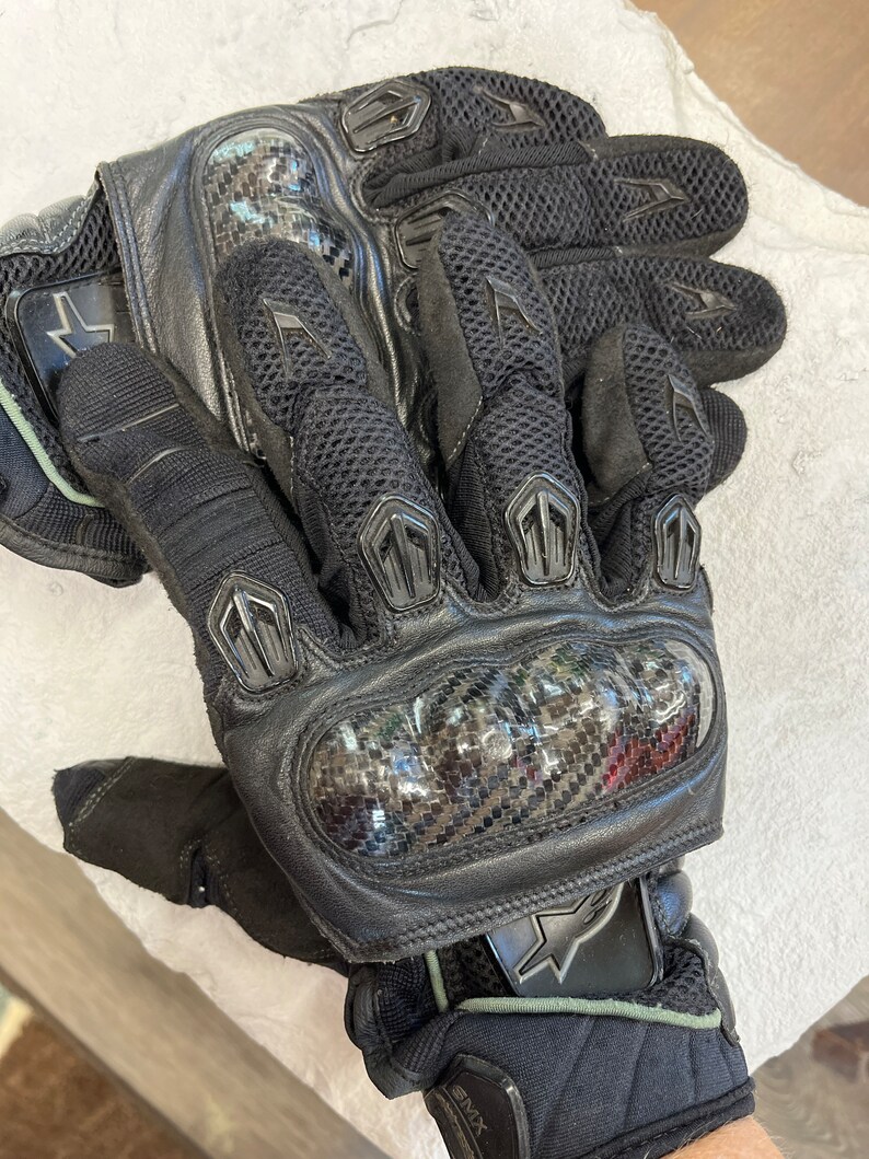 以下が含まれることがあります： カーボンファイバー製のナックルプロテクターとメッシュデザインを備えた、黒い革のオートバイ用グローブ。グローブは手首にベルクロ留めと星形のロゴが付いています。